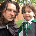 Jeff LaSala and his child are wearing cloaks held by leaf-shaped pins. Jeff has a steely gaze and trim beard while his child (who looks to be about eighteen months old) is pure grinning hobbit mischief.