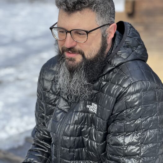 Shawn Gaffney, a scholar with salt-and-pepper hair and long beard, sits outdoors wearing horn-rimmed glasses, a puffy dark jacket, and a faraway gaze.