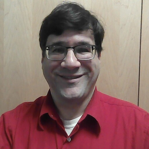This photo of David Trimboli shows his smiling face, horn-rimmed glasses, brown short hair unmarked by any grey. He is wearing a vivid red button down shirt and looks entirely approachable.