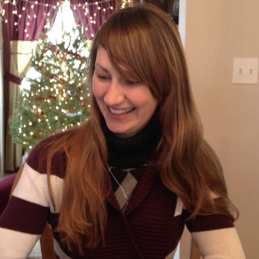 Eve Droma is smiling broadly looking down and sideways at something. Her reddish brown hair is long and flowy. There's a lighted tree in the background, and I think it's just darling that the maroon and cream colored curtains in the background match her color-block striped sweater.