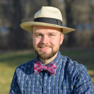 Keli Fancher is smiling directly at us, wearing his signature straw hat with black band, and a particularly jaunty fuchsia and blue bow tie. His blue shirt has pinstripes of printed edelweis chains and his green eyes and bearded smile somehow convey that he is a really fun dad!