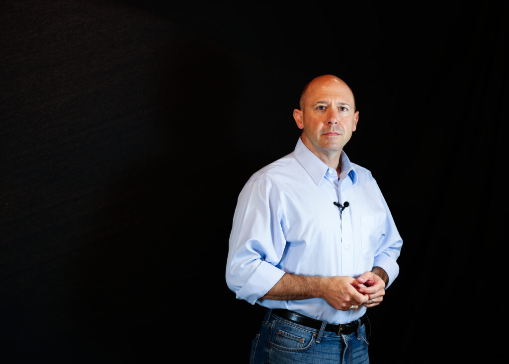 Mike Drout is looking right at the camera. His background is darkness but he is well lit. This three-quarter shot shows him in a comfortable situation, blue jeans with an ironed button-down shirt, the sleeves lightly rolled up. He's wearing a lavalier mike and his earnest gaze suggests that you are about to enter a marvelous wolrd of Old English knowledge