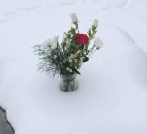 On a field of snow, white flowers in a vase with greens and a single red rose propre.