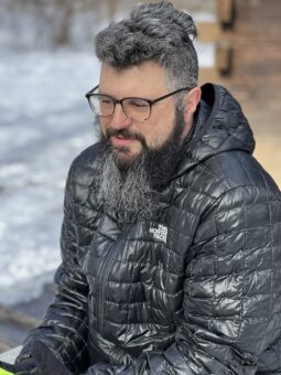 Shawn Gaffney, a scholar with salt-and-pepper hair and long beard sits outdoors wearing horn-rimmed glasses, a puffy dark jacket, and a faraway gaze.