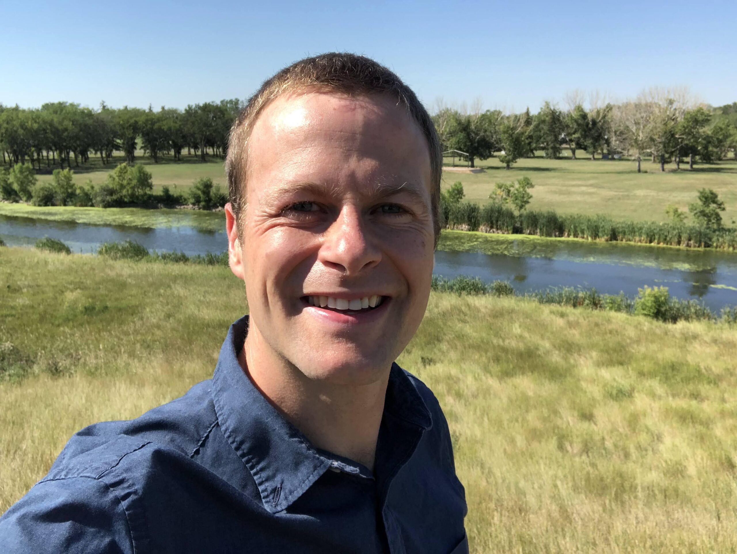 Rob Gosselin in his beloved great outdoors of Canada! He's grinning with real laugh lines around his eyes as though one or two of his many children are climbing up his legs while he's trying to take this selfie. The grasses are green behind him, the landscape includes a calm river and a dark far line of trees.