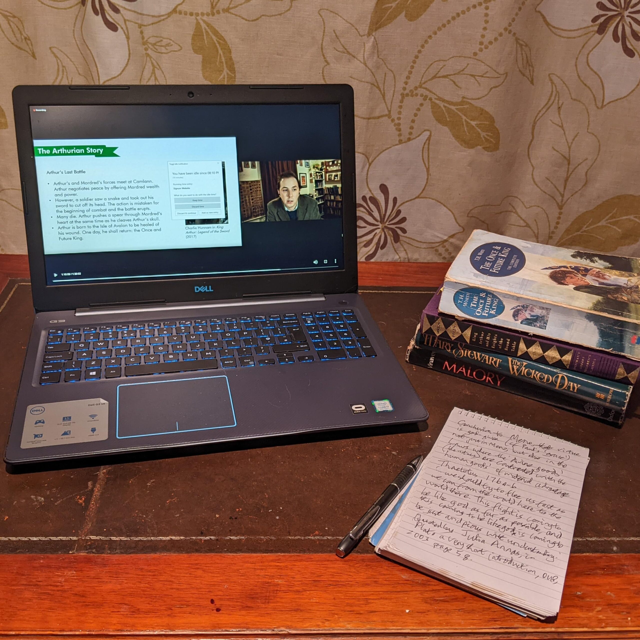 Decorative image of open laptop, stack of books icluding Malory's Morte D'Arthur, and a notebook and pen. The open laptop includes an image of Gabriel Schenk lecturing, as though the note-taker is jotting down something Dr. Schenk said in class to be used in the paper they are trying to write and very much need a Writer's Forge Wordsmith's advice on.