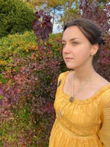A woman of European ancestry gazes thoughtfully into the distance. She stands by a hedgerow, clad in a very pretty yellow tunic or gown with an empire waist. Her long brown hair is clipped up and you wonder why she is so very fixedly staring behind you to your left... could there be... look out!