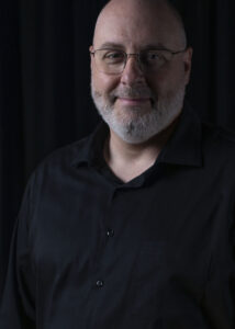 Corey Olsen's snow white beard and glasses might hint at his serious academic standing... but there's no way to hide that grin of delight, discovery and adventure. He's clad in dark colors before a dark background, but his eyes are shining.