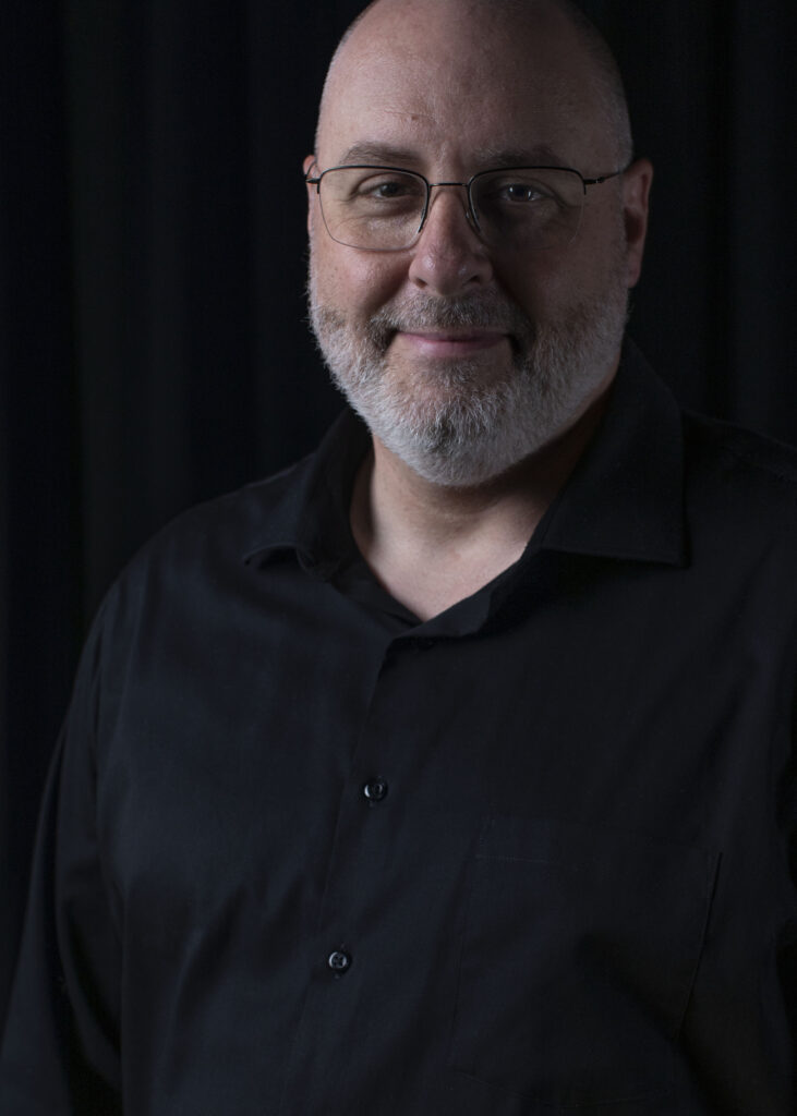Corey Olsen, wearing a black shirt and standing in front of a black curtain, cannot hide the light in his eyes—his enthusiasm for helping people to learn what they love best. Corey is a man of European descent with a trim salt-and-pepper bears, wire rimmed glasses, a gentle smile, and a tinkling eye.