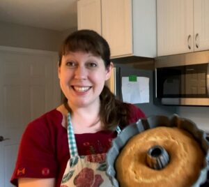 A warm and smiling woman offers a bundt-shaped seed cake fresh from the oven. Her bright eyes, gingham apron, and spotless kitchen tell us that this is a Mama on the go, leading the charge against chaos, one small child at a time.