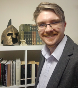 Julian Barr's bespectacled smile reveals a dimple or two in his neatly-trimmed red-blond goatee. He wears a crisp blue shirt and grey jacket and stands partly turned toward his collection of books—young paperbacks and venerable old hardbacks—which are punctuated by a wee replica statue of Lupa suckling Romulus and Remus and a replica bronze helm which looks like the one on the cover of the Chickering Beowulf,but given Dr. Barr's interest in rem Romanorum, it might be Roman, so I'll have to ask him about that.