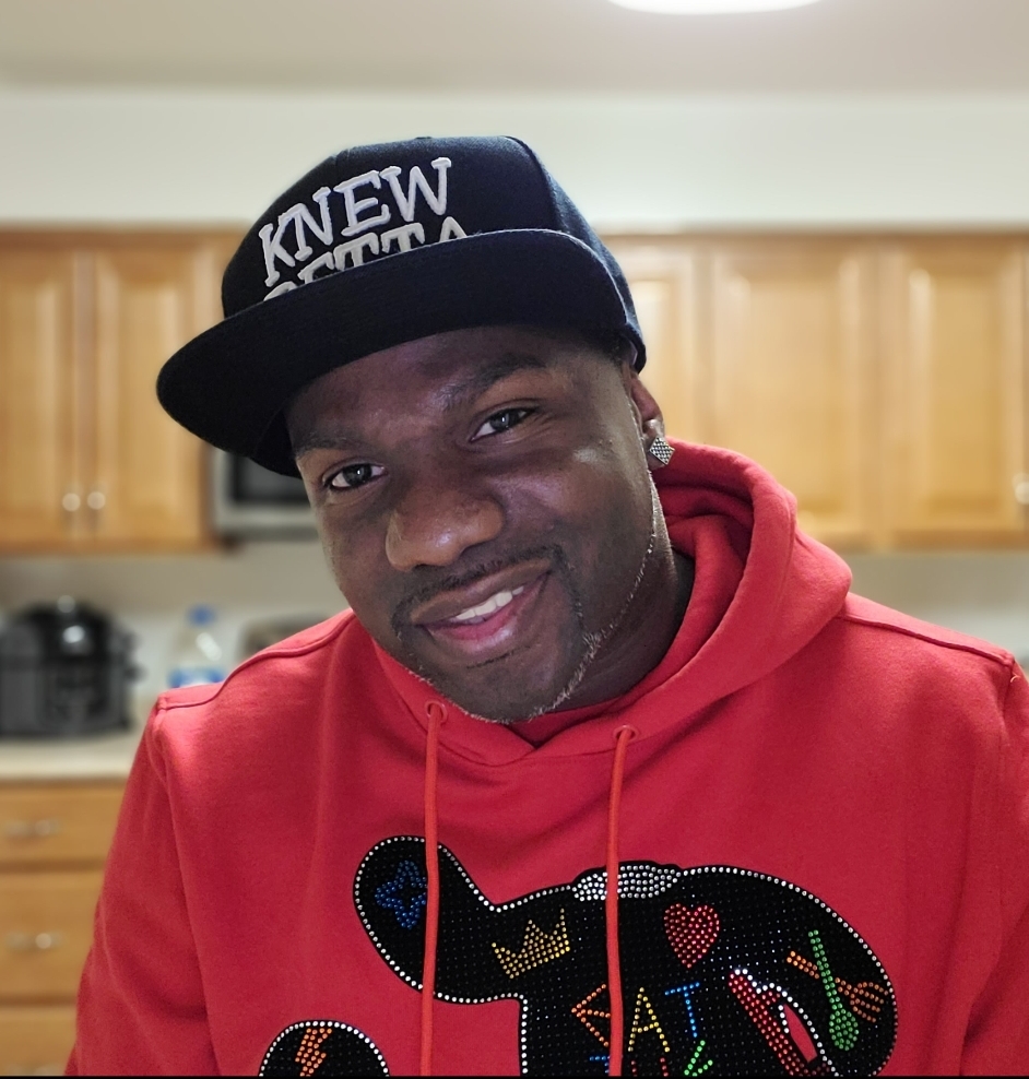 Knewbetta's warm smile invites us into the world of story. He's a man of African ancestry wearing a bright red cozy sweatshirt and a jaunty black baseball cap. He's standing in his kitchen with a gentle tilt of his head like he's welcoming us to sit down, kick up our feet, and listen to a great story.