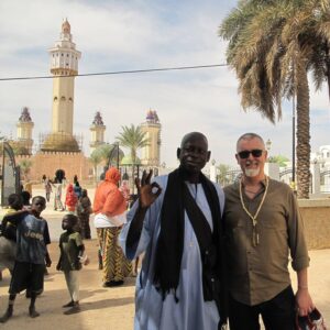 Patrick Malloy, a man of apparent European descent, visiting the beautiful city of Touba in Senegal. He smiles at us through white-bearded visage ad is walking with a friend through a beautiful outdoors with marvelous towers and a palm tree in the distance.
