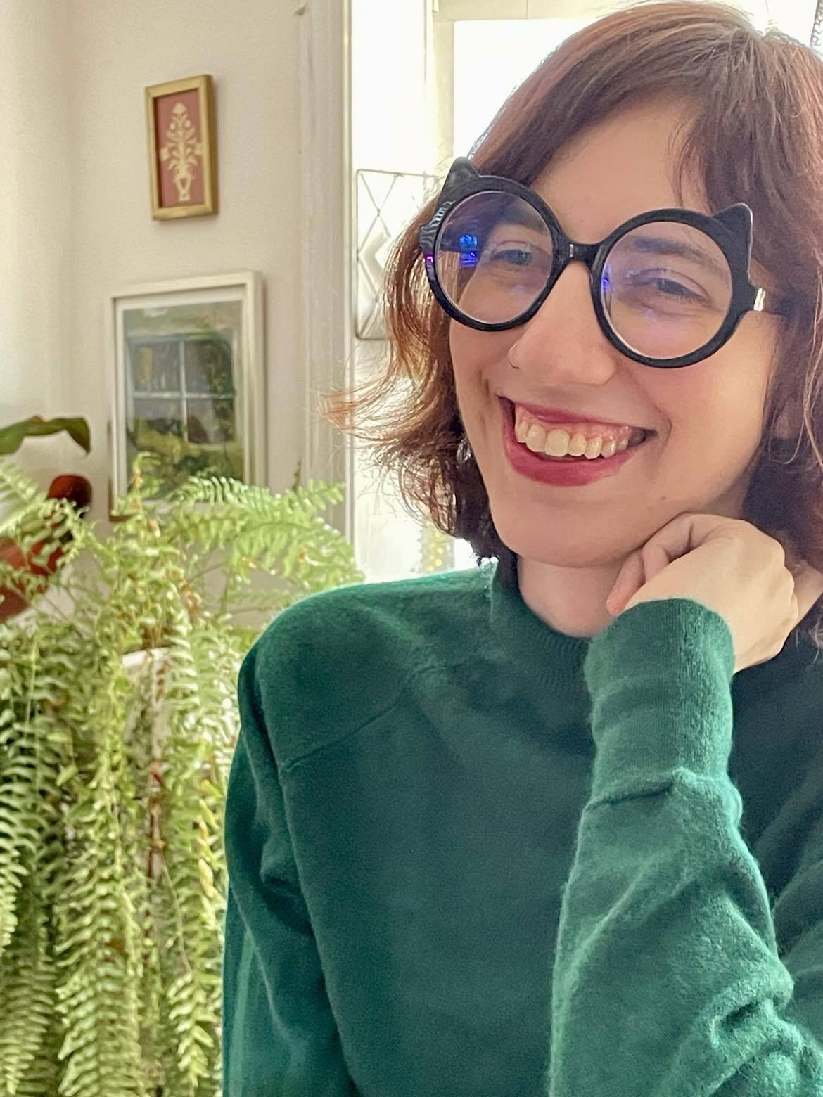 This lovely woman of european ancestry wears cat-rimmed glasses and a huge smile. She is engaged in conversation with a rather large indoor fern on the topic of plant representation in anime.