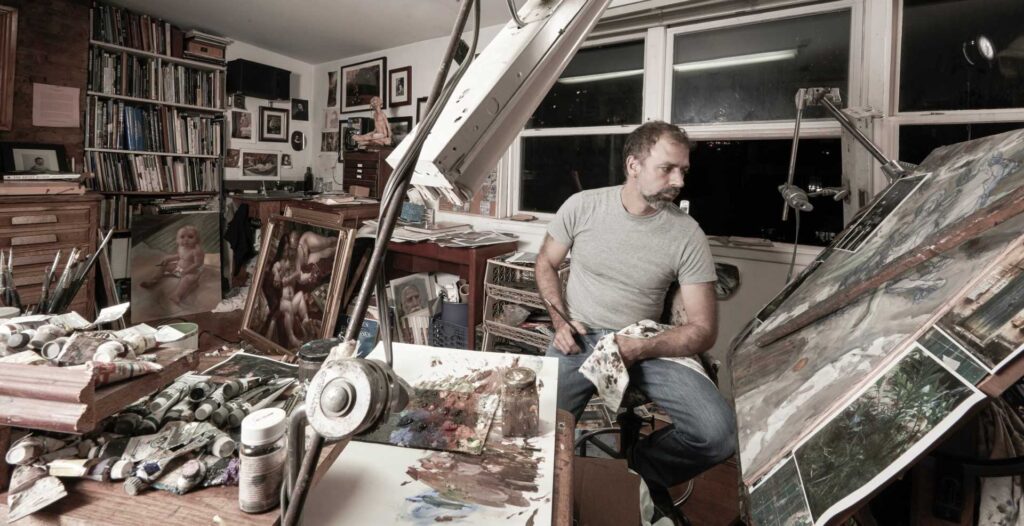 Artist Donato Giancolo sits in his studio in a grey t-shirt and jeans surrounded by canvases both finished and unfinished.