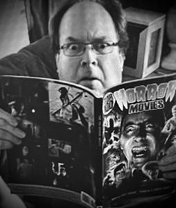 A bespectacled reader with eyes wide and brows furrowed is reading a magazine called "Horror Movies."