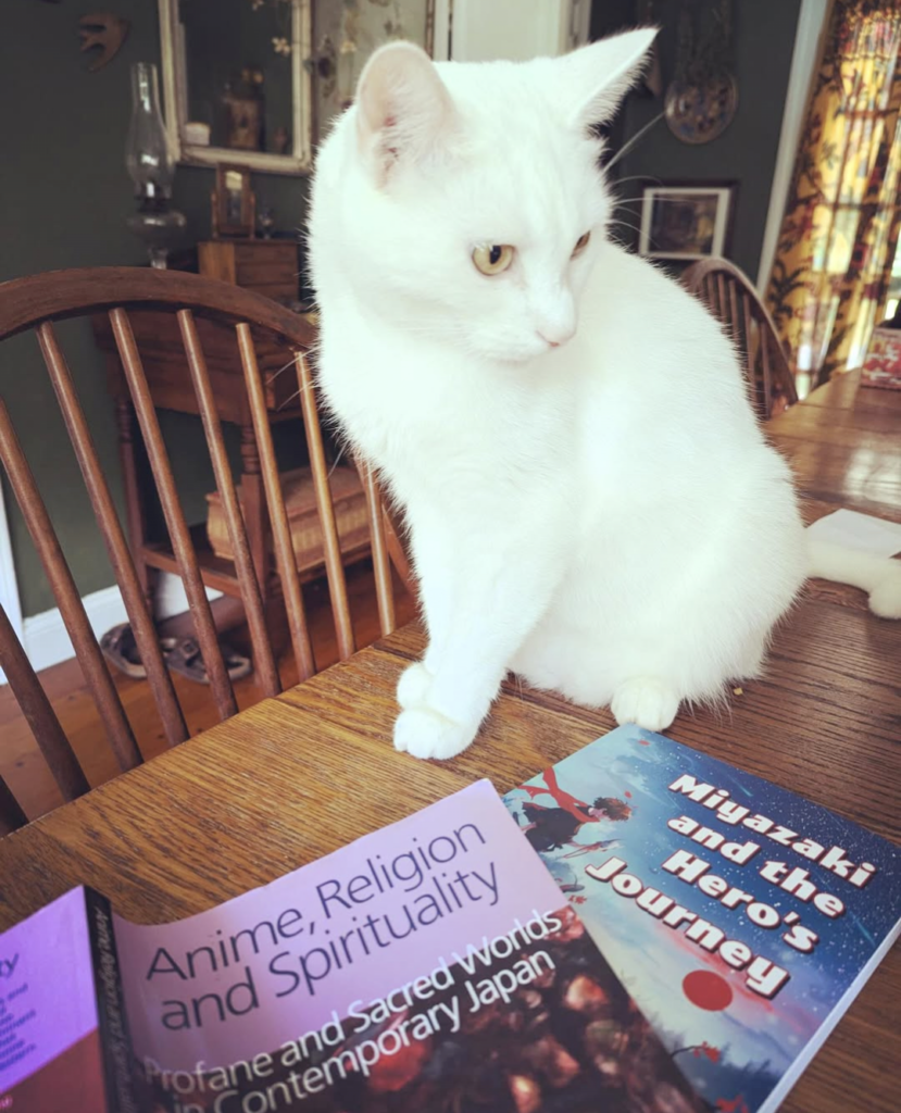 Pangur Ban, a beautiful white cat, is casually looking at the book _Miyazaki and the Hero's Journey_