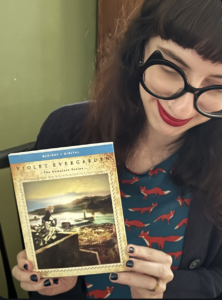 To no one's surprise, Bronwyn is holding a book! She is a woman with dark hair, cat-eye glasses, and a broad grin. She is in fact charged by touching books, similarly to the way plants make energy from touching light. Here eyes are totally focused on the copy of Violet Evergarden in her hands!