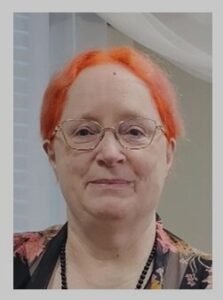 Nancy Raines embraces life as a cheerfully-carrot-topped redheaded, bespectacled, grinning person with really colorful autumn-leaves colored jacket in this headshot.