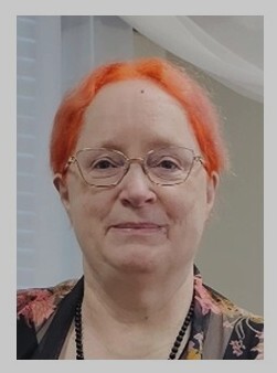 Nancy Raines embraces life as a cheerfully-carrot-topped redheaded, bespectacled, grinning person with really colorful autumn-leaves colored jacket in this headshot.