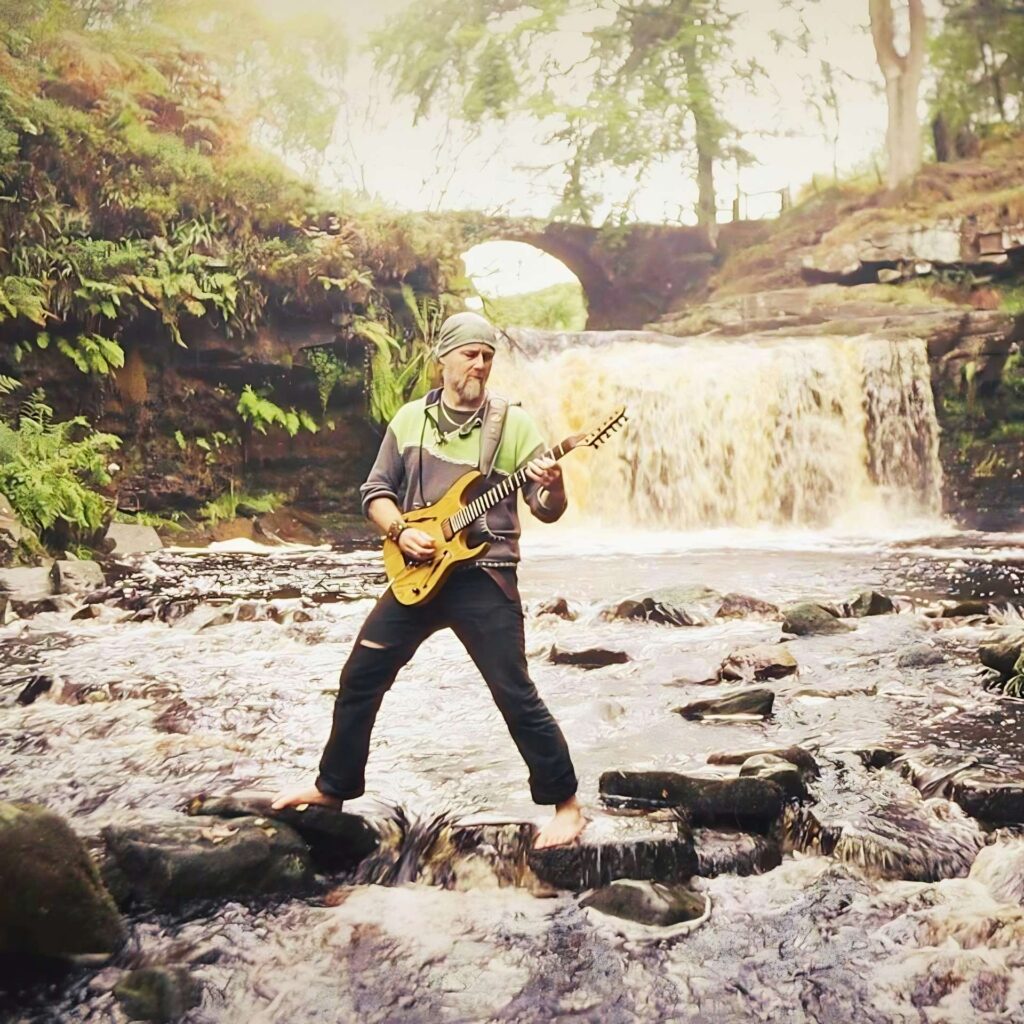 Dave Brons, musician, playing an electric guitar while standing on some rocks in the middle of a river. There is a waterfall behind him.