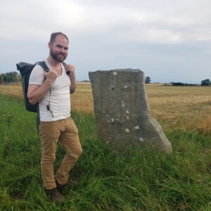 Galen's got a backpack, a beard, and a fabulous hiking attitude! He's standing next to a very old, weathered rock with actual runes on it, which is the coolest thing ever!