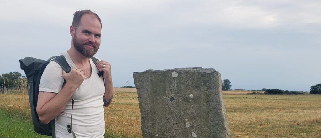 Galen's got a backpack, a beard, and a fabulous hiking attitude! He's standing next to a very old, weathered rock with actual runes on it, which is the coolest thing ever!